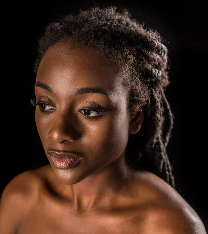 Woman with a natural hair style of full locs stands in a studio against a black background while appearing to not wear a shirt