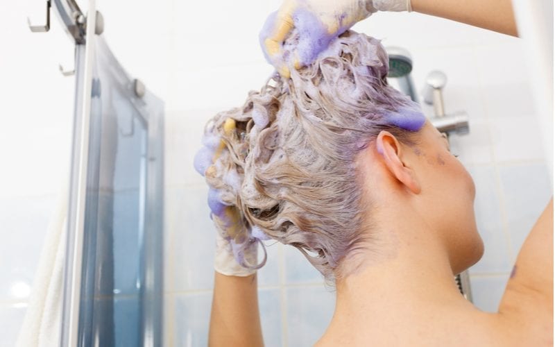 Woman washing hair toner out of her hair in a shower that has purple shampoo in it