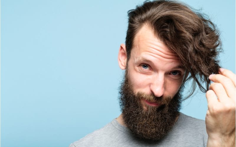 Against a blue background stands a guy with a side-swept top and he holds his long bangs while rocking a beard