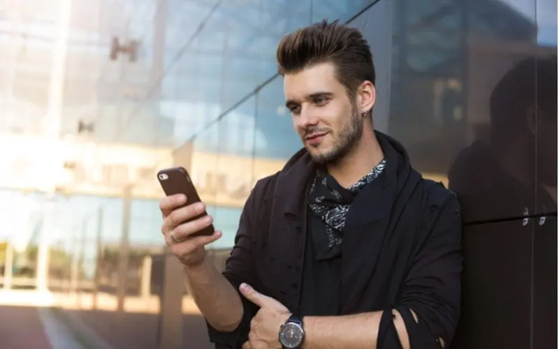 Bearded man with an up-and-back hairstyle for round faced men looks at his phone while leaning against a glass wall