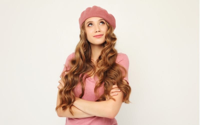 Woman in a French looking hat with a long curly brown weave and a pink shirt looks up while crossing her arms