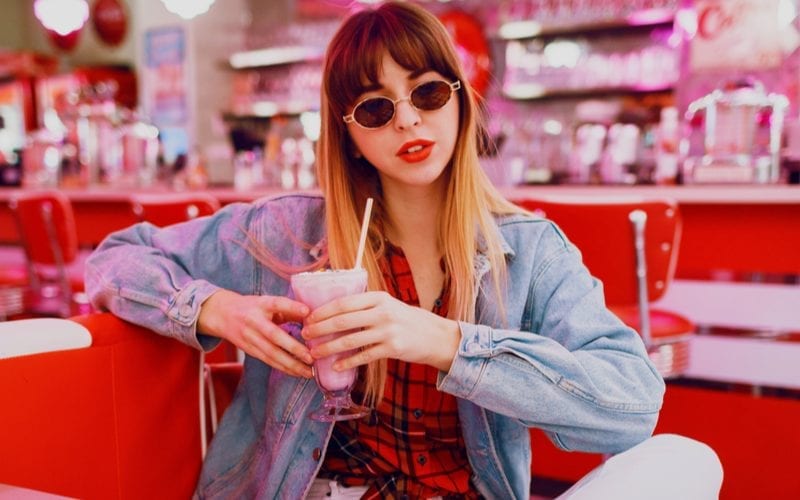 Fashionable woman in tan jeans sits cross legged in an ice cream parlour while wearing a jean jacket, red lip, and curtain bangs