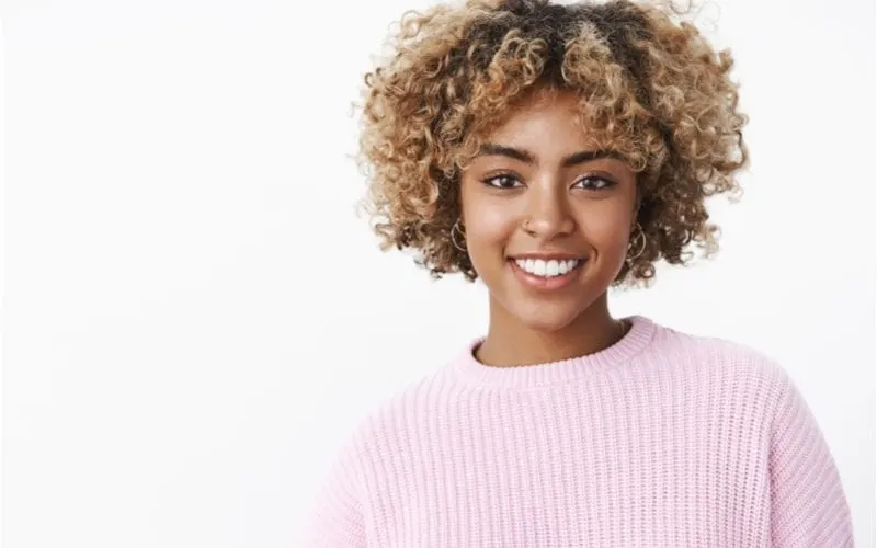 Ethnic woman in a white ribbed sweater smiles and wears natural curly curtain bangs