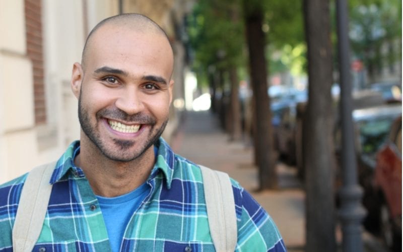 Man who is shaved with a beard smiles and wears a backpack while standing outside a tree-lined sidewalk
