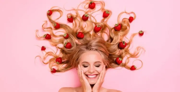 Woman with strawberry blonde hair literally with the fruit in her hair lying on a pink background and holding her smiling face