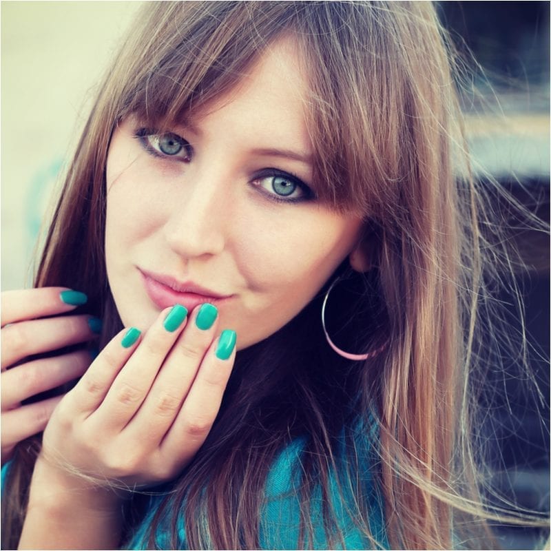 Lady with green nails holds her chin and her curtain bangs frame her pretty blue eyes