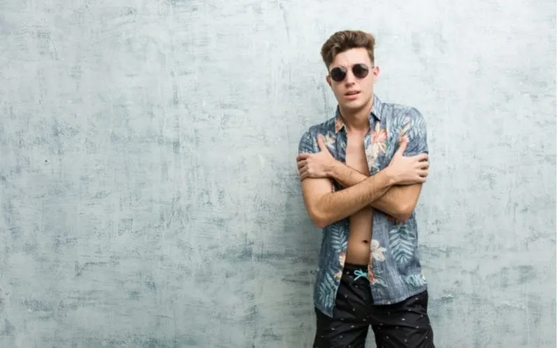 Guy with an unbuttoned floral shirt stands against a concrete-looking studio backdrop while wearing sunglasses and crossing his arms for a piece on hairstyles for wavy haired men