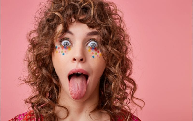 Strange portrait of a woman with curly curtain bangs wears polka dots on her cheeks and sticks her tongue out while opening her eyes up wide