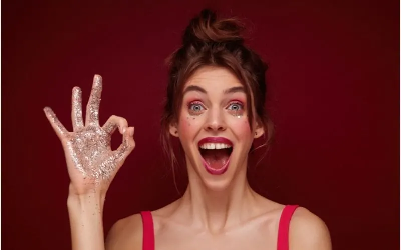 Obnoxious image of a lady with a messy half-up bun with glitter on her hand and curtain bangs giving the OK sign