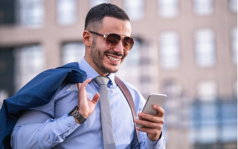 Smiling man with a fade buzz cut looks at his phone and holds his suit jacket above his right shoulder
