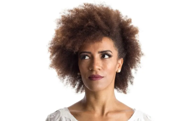 Woman with a natural pinned afro hairstyle looks up to her right while standing in a bright room and wearing a frilled white shirt