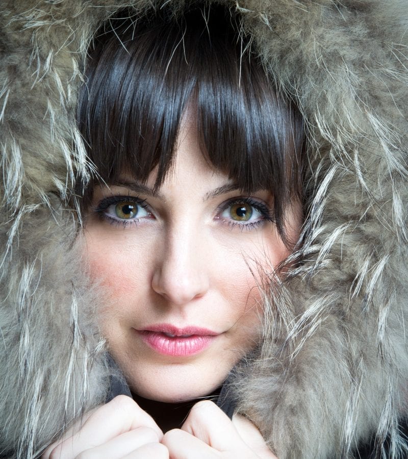European looking woman in a furry hooded jacket holds the hood at its ends and looks intensely at the camera without smiling