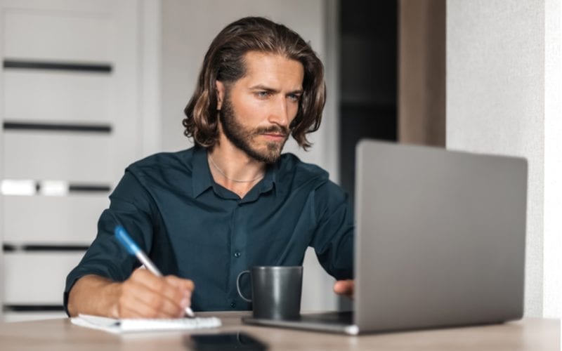 Man with long hair holds a pen in his right hand and touches a key on his laptop which sits on a desk next to his coffee
