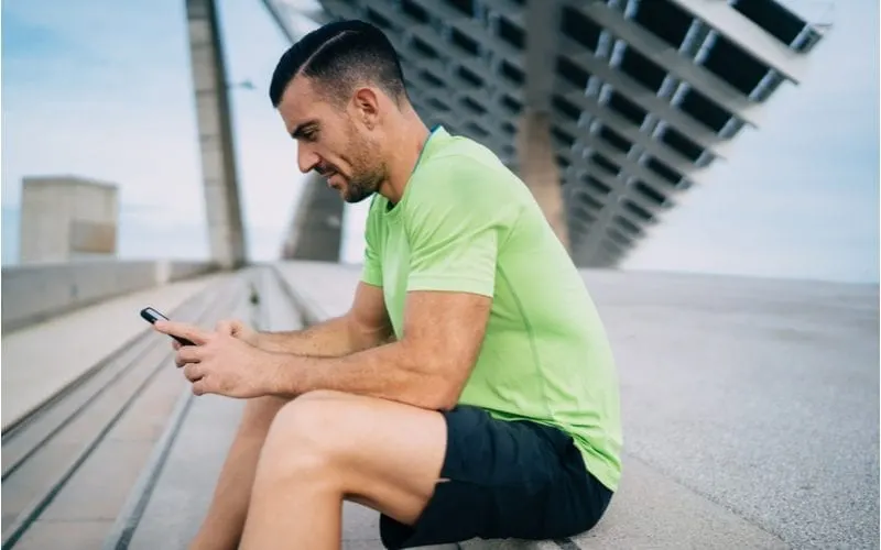 Man dressed in athletic clothing and a high drop fade with shaved part looks at his phone while sitting on a concrete step