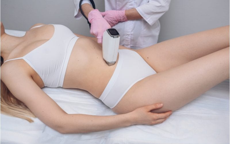 For a piece on how much is laser hair removal, a woman laying on her back in a white sports bra and panties gets the treatment done on her lower stomach