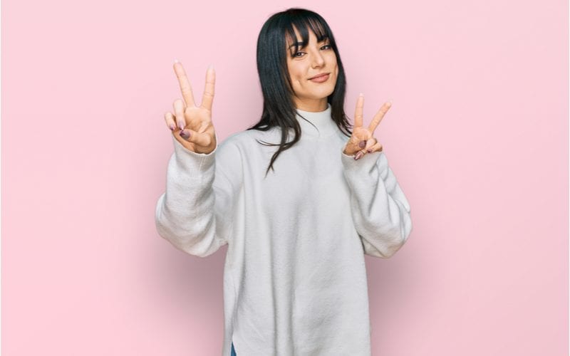 Lady with short curtain bangs and a flowing grey shirt gives the peace sign with both hands and grins with large dimples