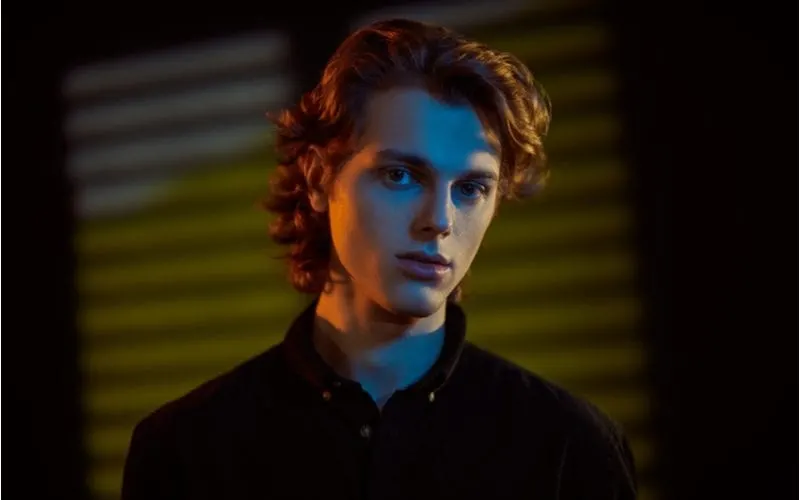 Handsome young man with wavy hair stands in a dark room with slightly open blinds in the background