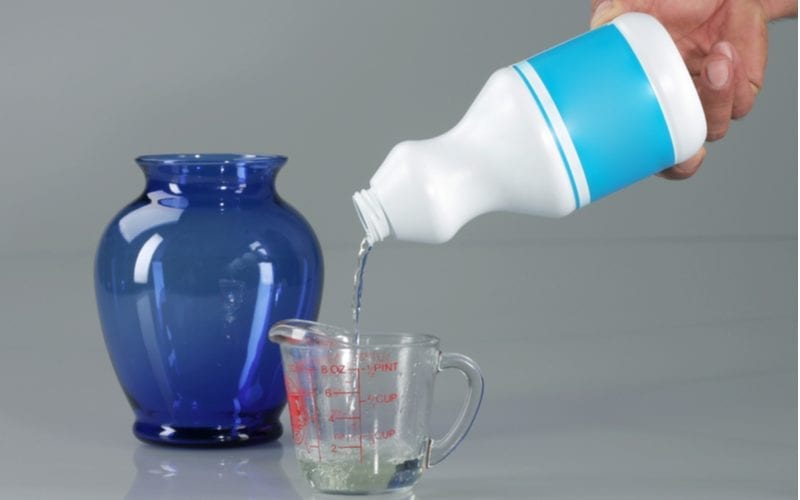 Hand pouring bleach into a measuring cup that sits in front of a glass bowl or vase for a piece on how to remove hair dye
