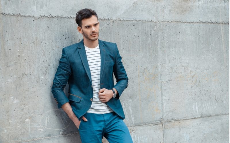 Man in green pants and a short teal blazer leans against a concrete wall rocking a hairstyle for wavy hair men