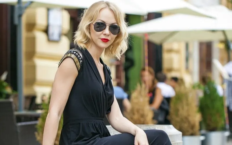 Young thin woman in a one-piece full-legged black jumper sits on a modern concrete and wooden bench and wears aviators with curtain bangs