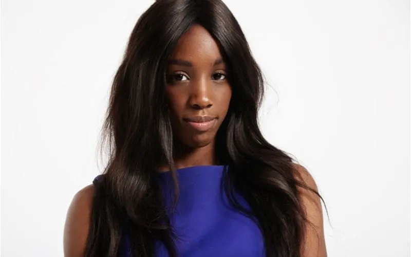 Young woman in a pretty blue dress and a long weave hairstyle stands in a studio with a white background without smiling