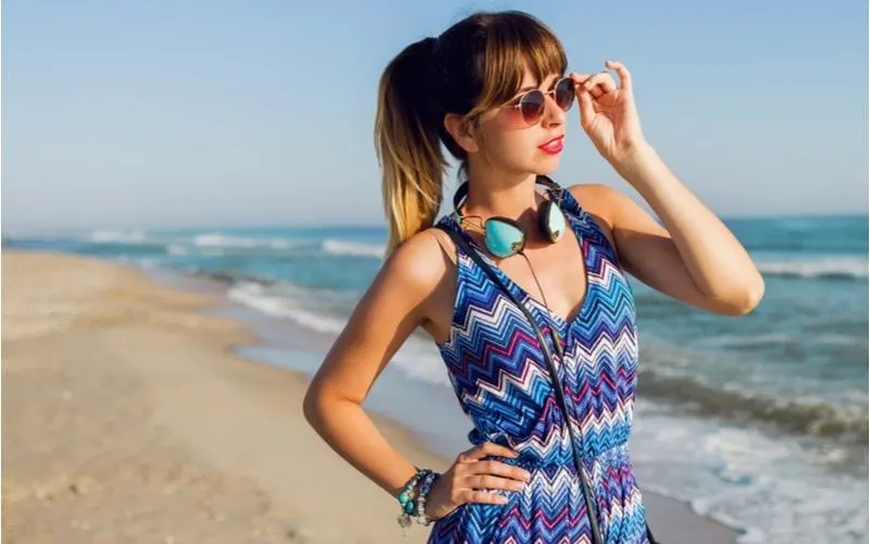 Hipster woman with dark round glasses and teal headphones holds her right hip and stands in front of an ocean