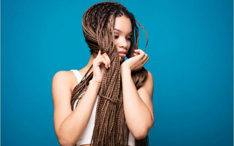 Young woman with mini box braids wears a cutoff midriff shirt and holds her hands up to her face while standing in front of a blue background