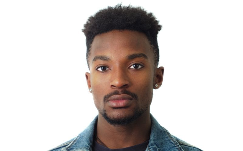 Clean cut black man with diamond earrings looks at the camera against a white background in a studio and wears a jean jacket