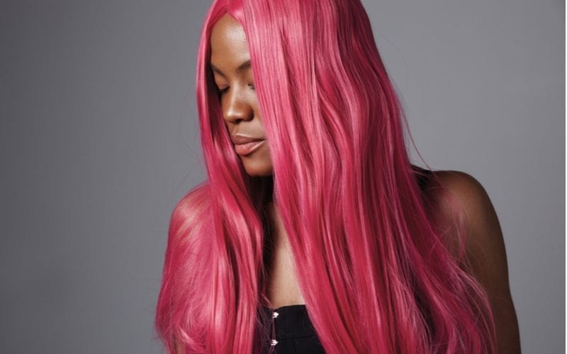 Black woman with shiny long pink weave hairstyle closes her eyes and looks right while standing against a grey background