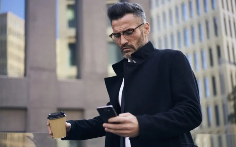 Man in a pea coat holds a coffee in his right hand and a phone in his left while walking outside a pair of high-rise buildings