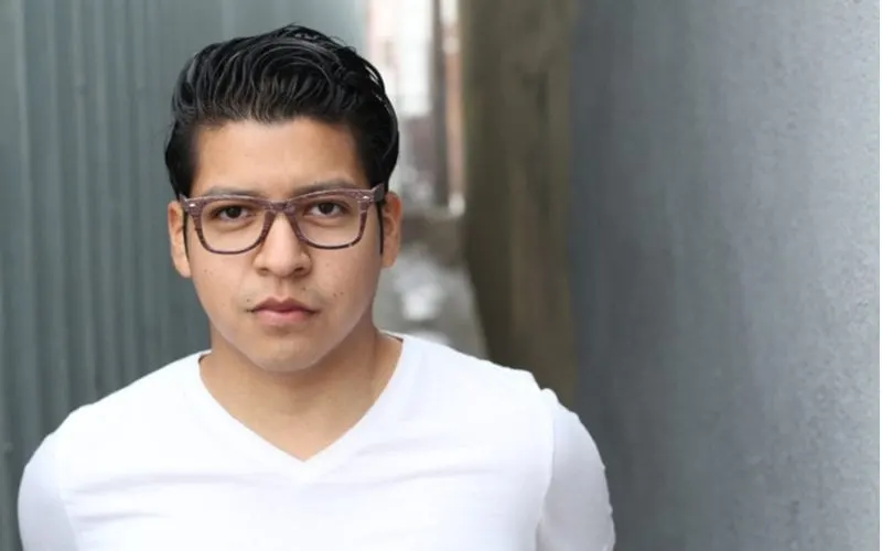 For a piece on haircuts for round faces (men), a guy with a long crew cut and a white v neck in glasses looks at the camera while standing in an alley