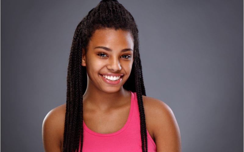 Lady with half-up box braids and a pink shirt stands in front of a gray background in a studio and smiles