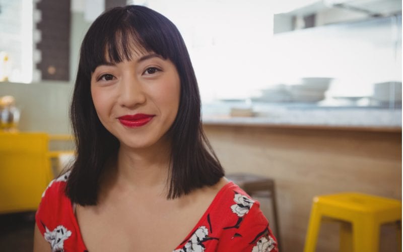 Pretty asian woman with straight-across bangs in a red floral shirt stands in front of a modern kitchen