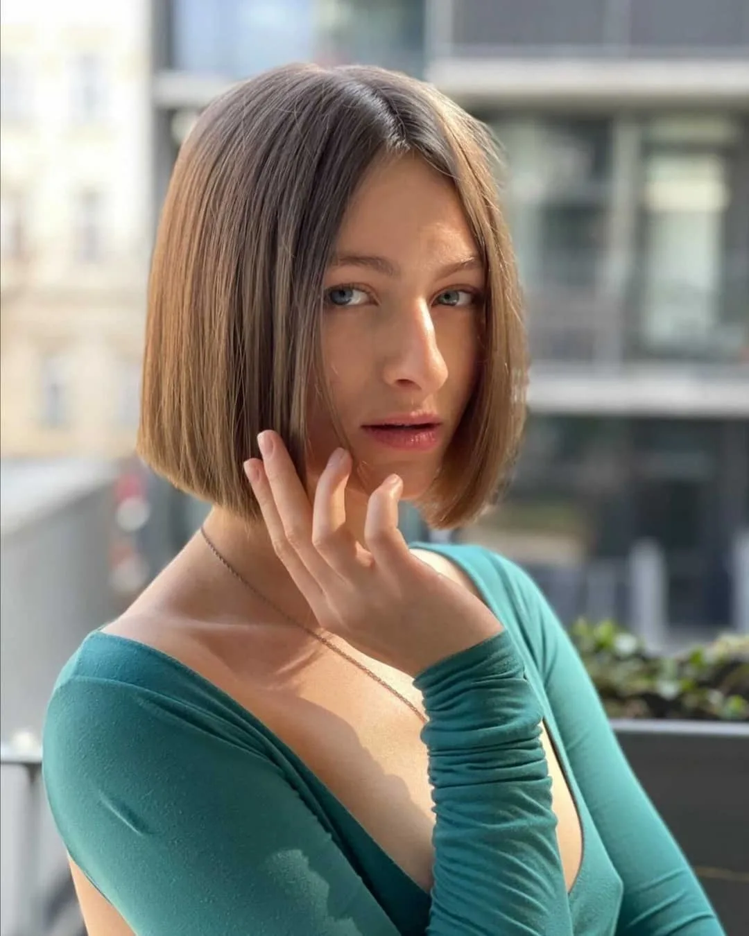 Petite Russian woman in a green open-back shirt stands on a balcony showing off her new bob haircut that is ash brown in color