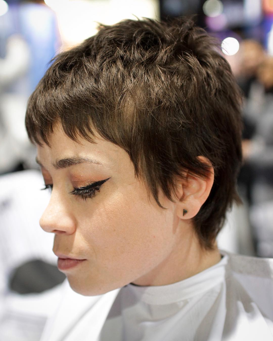 Lady with an ash brown pixie hair cut sits solemnly in a cape with dark eyeliner 