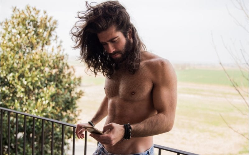 Sexy guy without a tshirt standing on a balcony in front of a grassy lawn lets his long hair blow in the wind