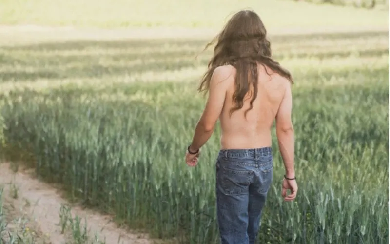 Man with long hair and mid-back waves walks shirtless in blue jeans through a field of high grass