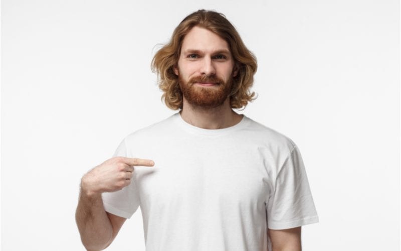 Man in a white tshirt points at his right pec while wearing a long hairstyle for men and standing in a white room