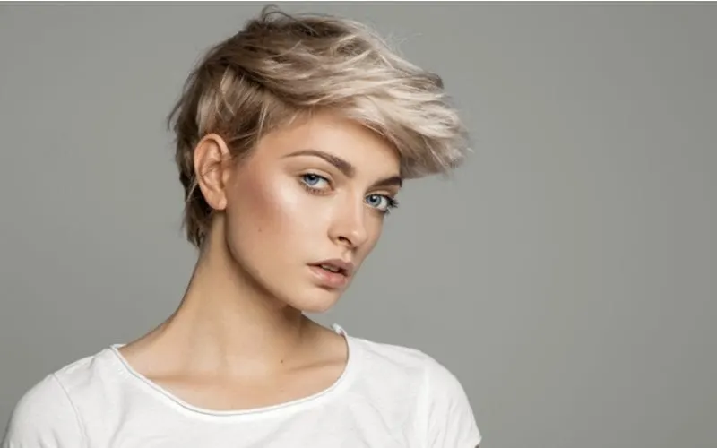 As a featured short hairstyle for a square faced woman, a lady in a white crew neck shirt wears a pixie cut and looks out of the corner of her eye