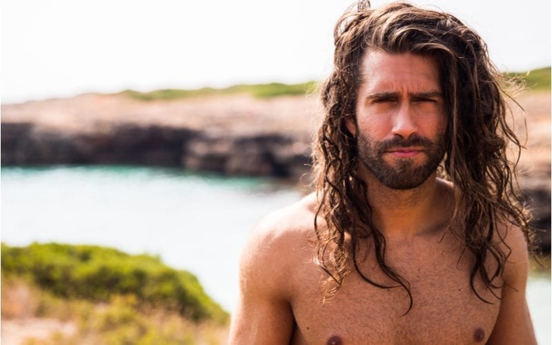 Man with a long Castaway-style haircut and no shirt standing in front of a stream