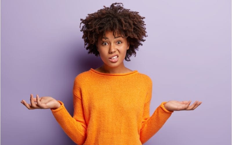 For a piece on what women's haircut should I get, a woman with an afro and an orange sweater holds her arms up in confusion