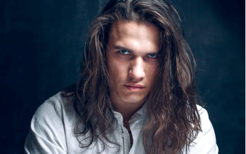 muscular man in a rough white button up shirt and a long hairstyle for men looks intently at the reader