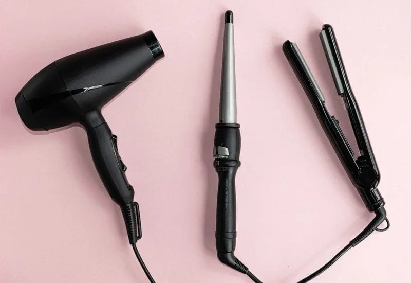 On a pink background sits a lay flat image of a black hair straightener, hair dryer, and a curling iron