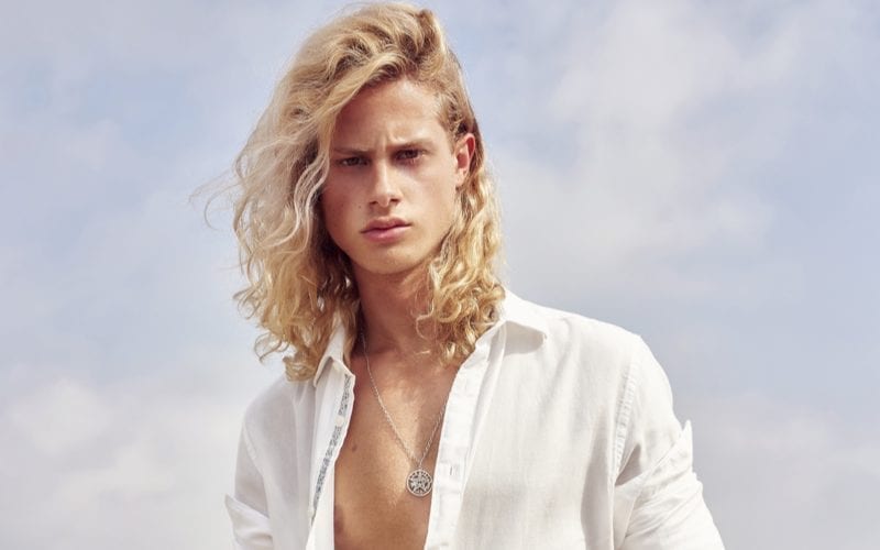 For a piece on men's long hairstyles, a guy in a white shirt that is unbuttoned looks sternly at the camera and stands in front of a cloud-dotted sky