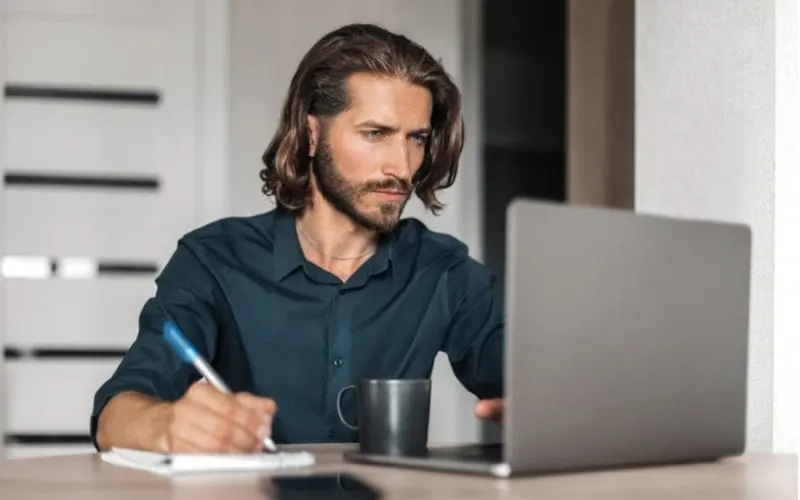 Guy with a long haircut for men writes on a tablet of paper while looking at a laptop