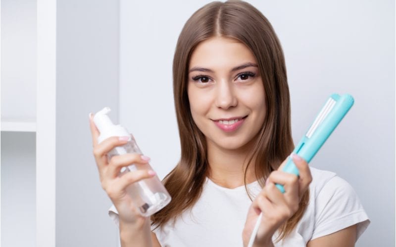 lady in a salon with shiny hair holding a bottle of the best heat protectant and another holding a teal flat iron standing in front of a bookshelf