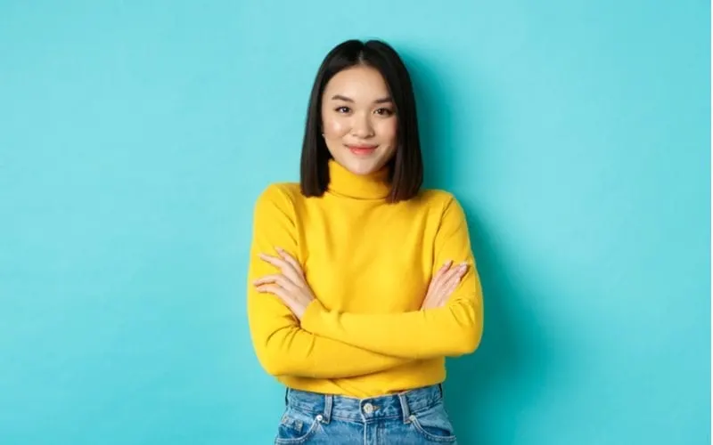 Asian woman in a yellow sweater stands crossing her arms and looking at the camera in a blue room