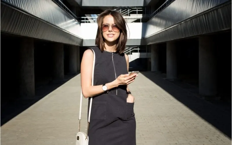 Lady in a black courtyard in a black dress with dark medium-length hair puts her hand in her pocket and looks at her phone