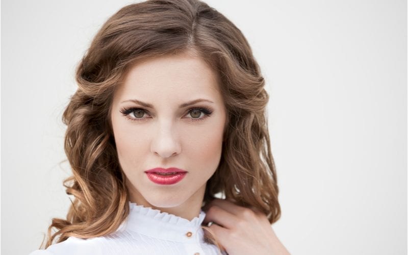 Romantic young woman stands seductively holding the end of her ash brown hair while wearing red lip and very heavy upper eye makeup