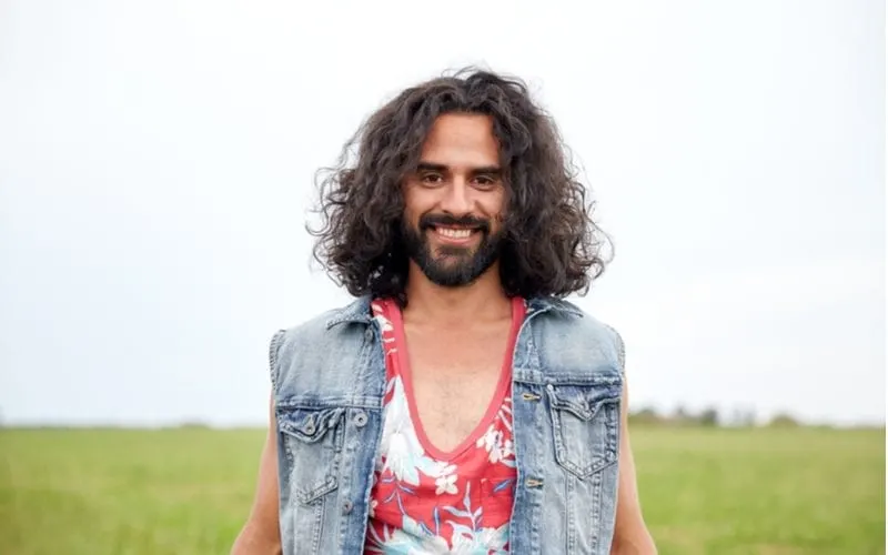 For a roundup on long hairstyles for men, a Russell Brand lookalike wears a flowery tank top and a jean vest and smiles big in a cornfield
